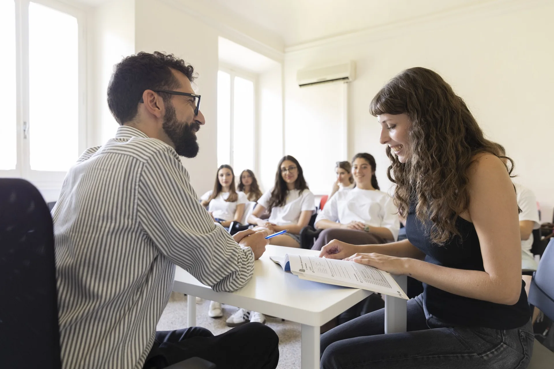 A teacher and a student at a table with an open book, the class behind them