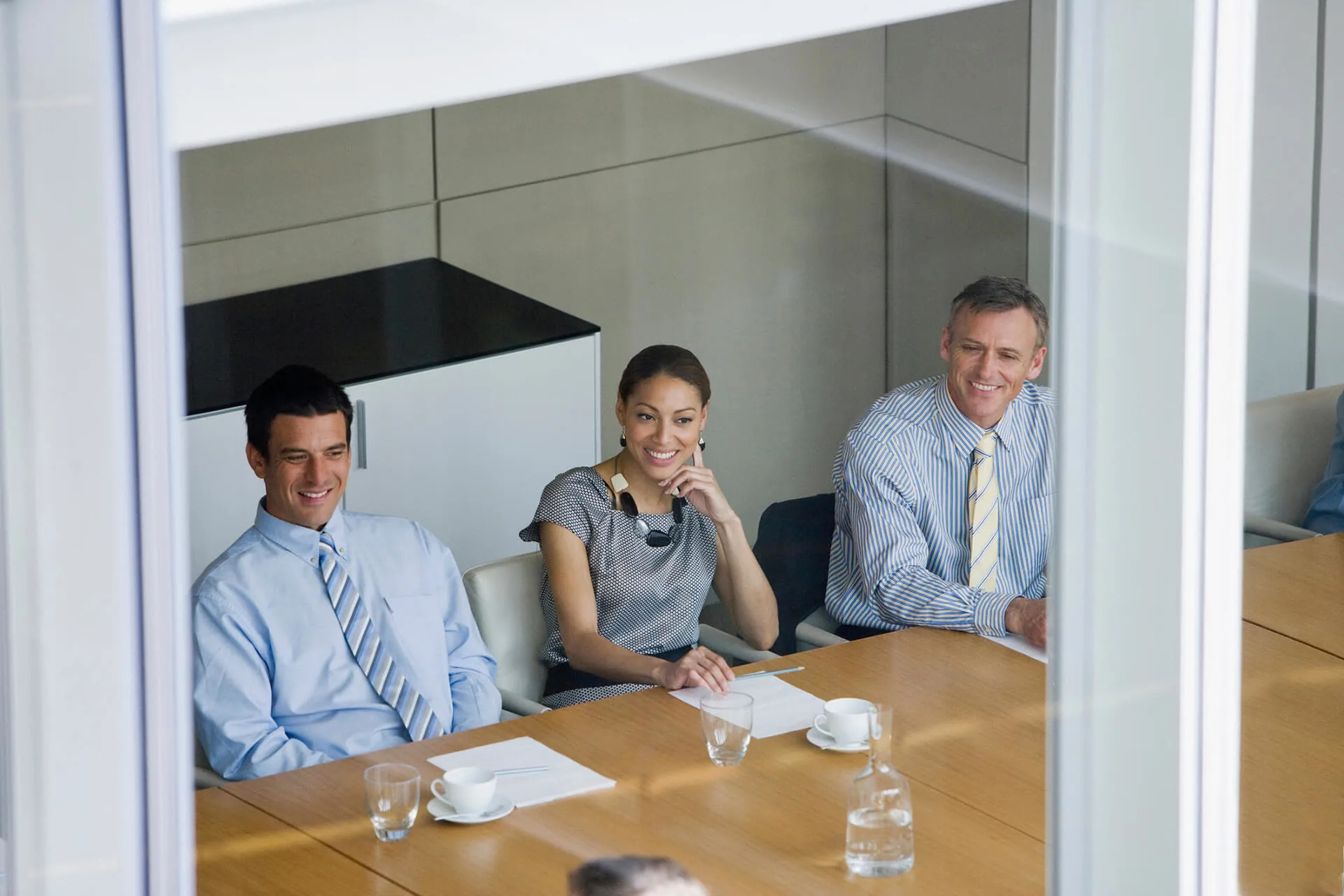 Three professionals smiling during a meeting, seen through the glass wall of a meeting room